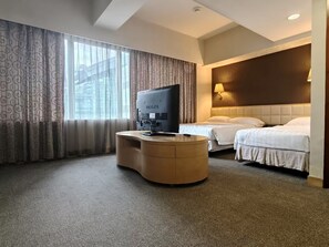 Ein Hotelzimmer mit einer gebogenen Schreibtisch, einem Fernseher, zwei Betten und einem großen Fenster mit Vorhängen.
