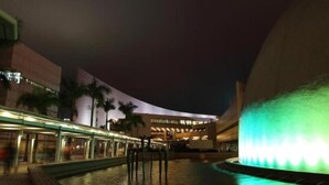 A nighttime view of a modern building with a curved facade, a water feature with green lighting, and palm trees in the background. - H1 hotel (Kowloon)