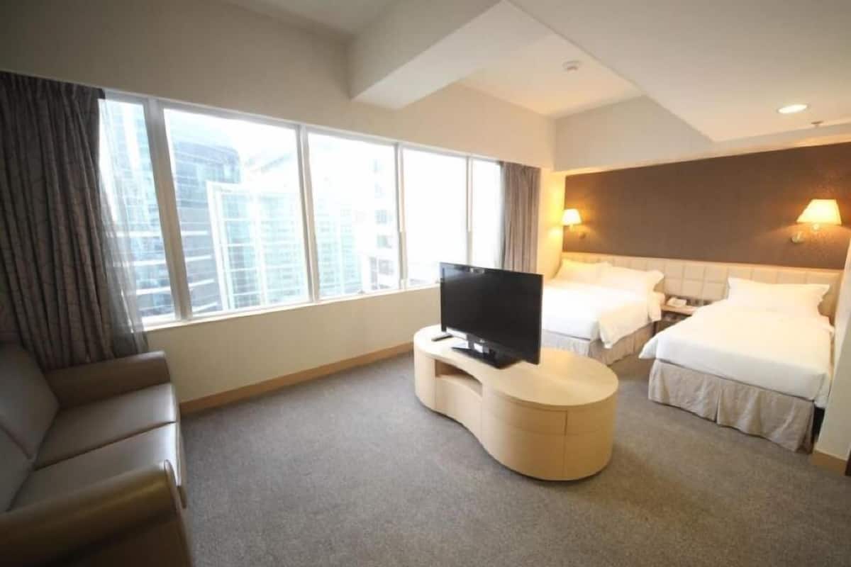 A hotel room with a bed, a TV on a stand, a sofa, and large windows offering a city view.