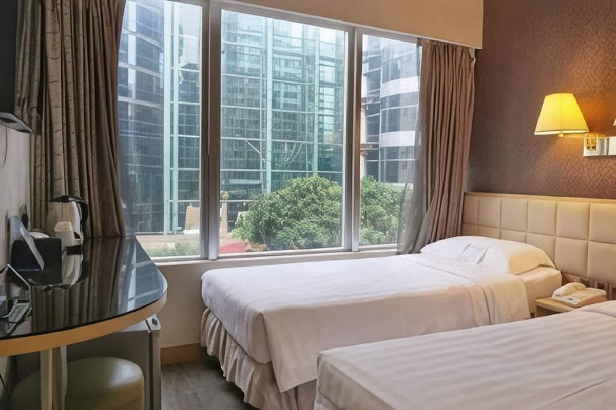 A hotel room with two beds, a desk, and a large window offering a view of buildings and greenery.