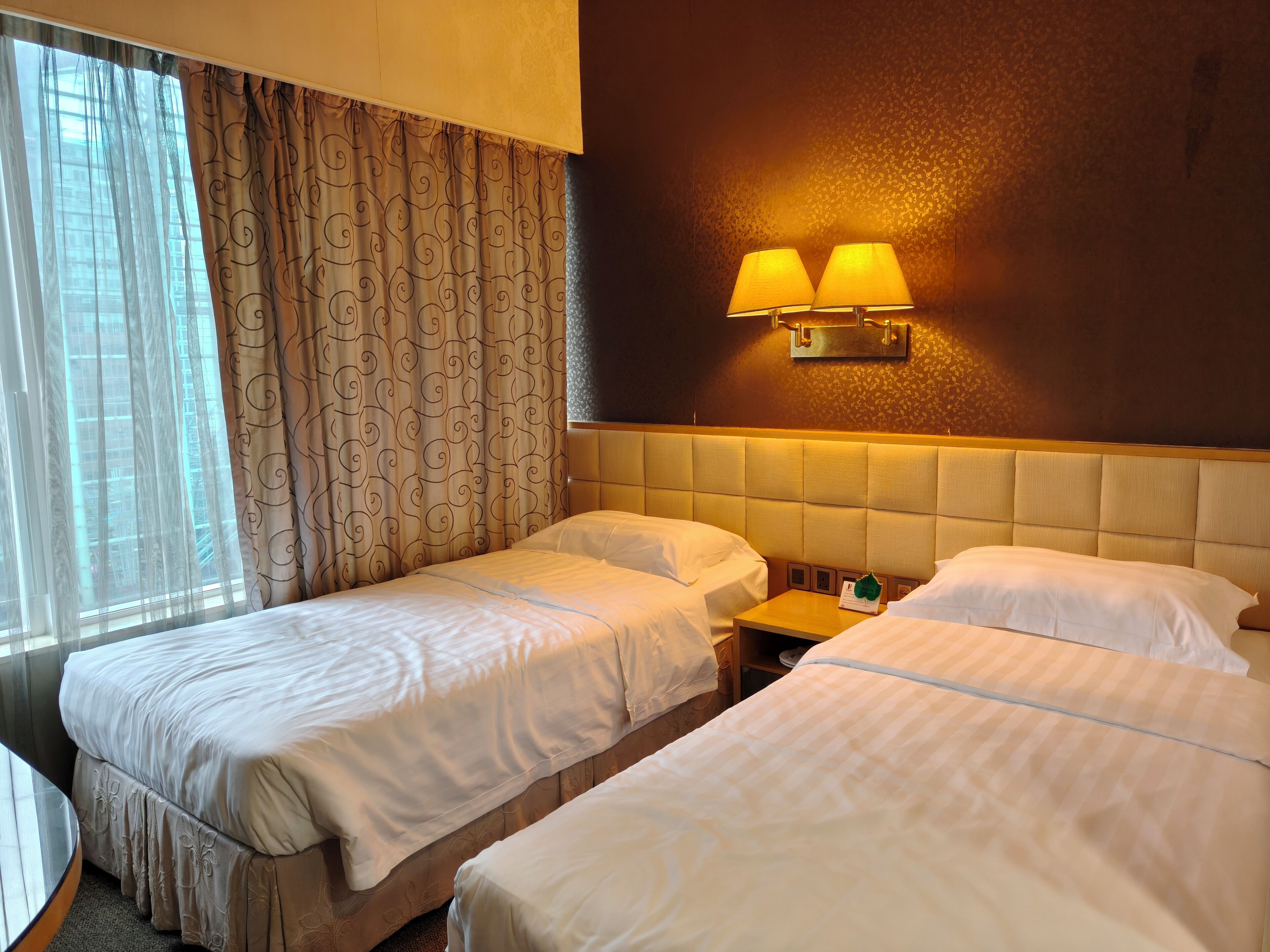 A hotel room with two beds, a small table, and a wall-mounted lamp.