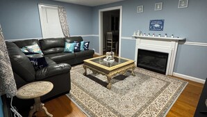 Uma sala de estar com um sofá de couro preto, uma mesa de centro de vidro, um tapete estampado, uma lareira e uma parede azul.
