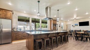 A modern kitchen with a large island, bar stools, wooden cabinets, stainless steel appliances, and pendant lighting.