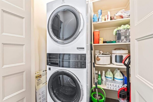 Una lavadora y secadora apiladas en un cuarto de lavado, con estantes superiores que contienen productos de limpieza y una escoba.