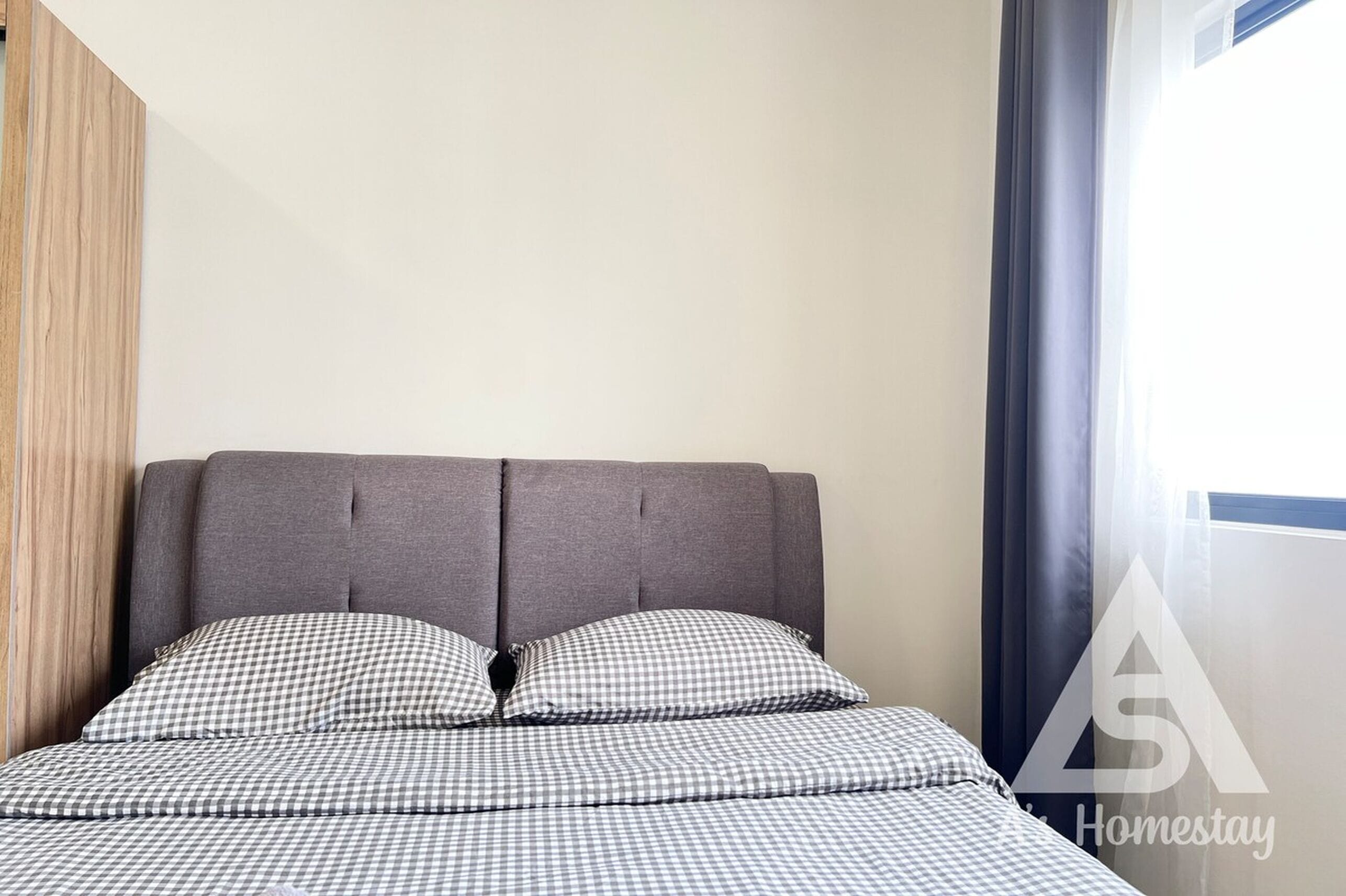 A neatly made bed with checkered bedding, a grey upholstered headboard, and a window with curtains.