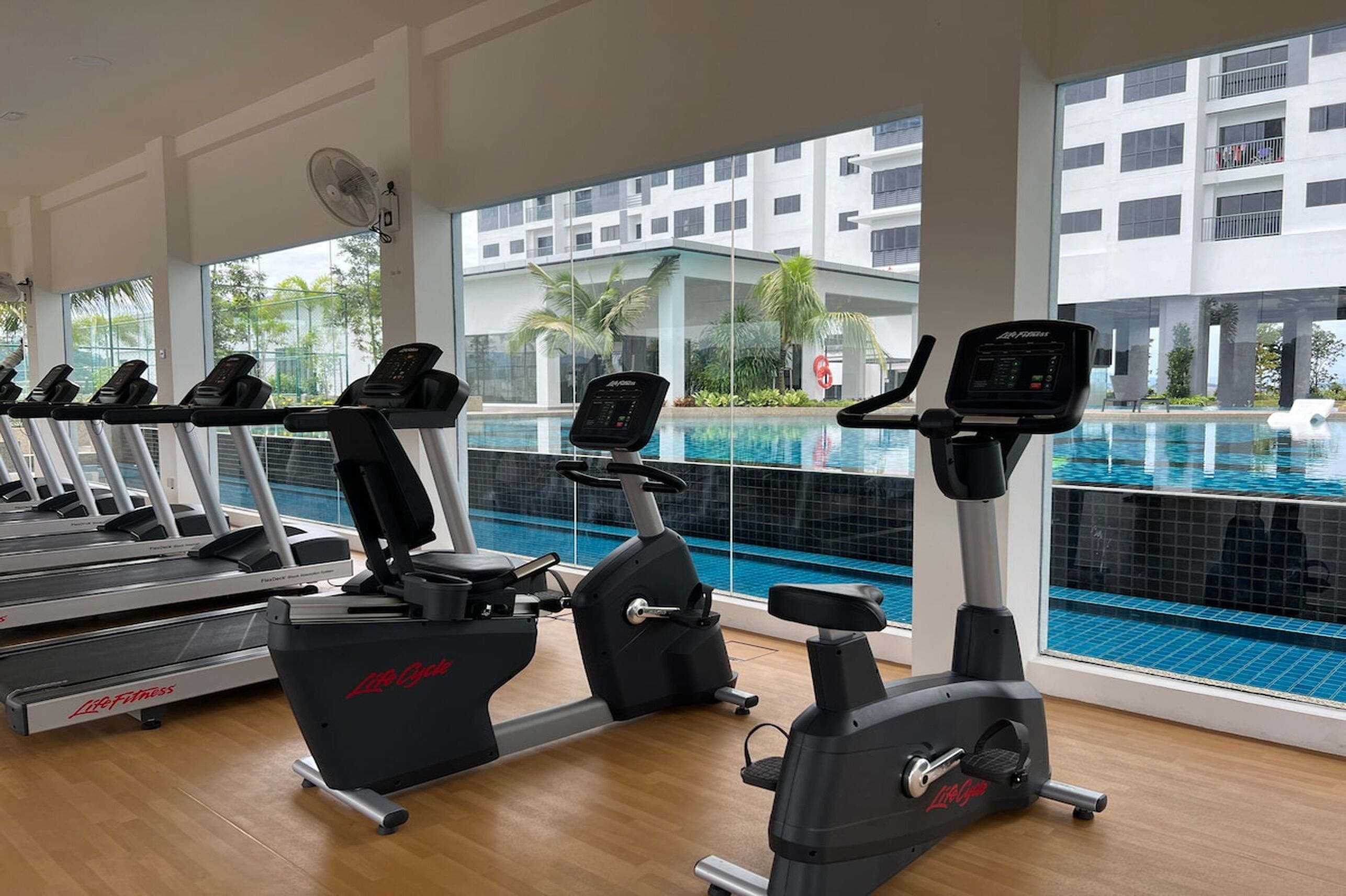 A gym with treadmills, stationary bikes, and an elliptical machine. It has large windows with a view of a swimming pool and a building with balconies.