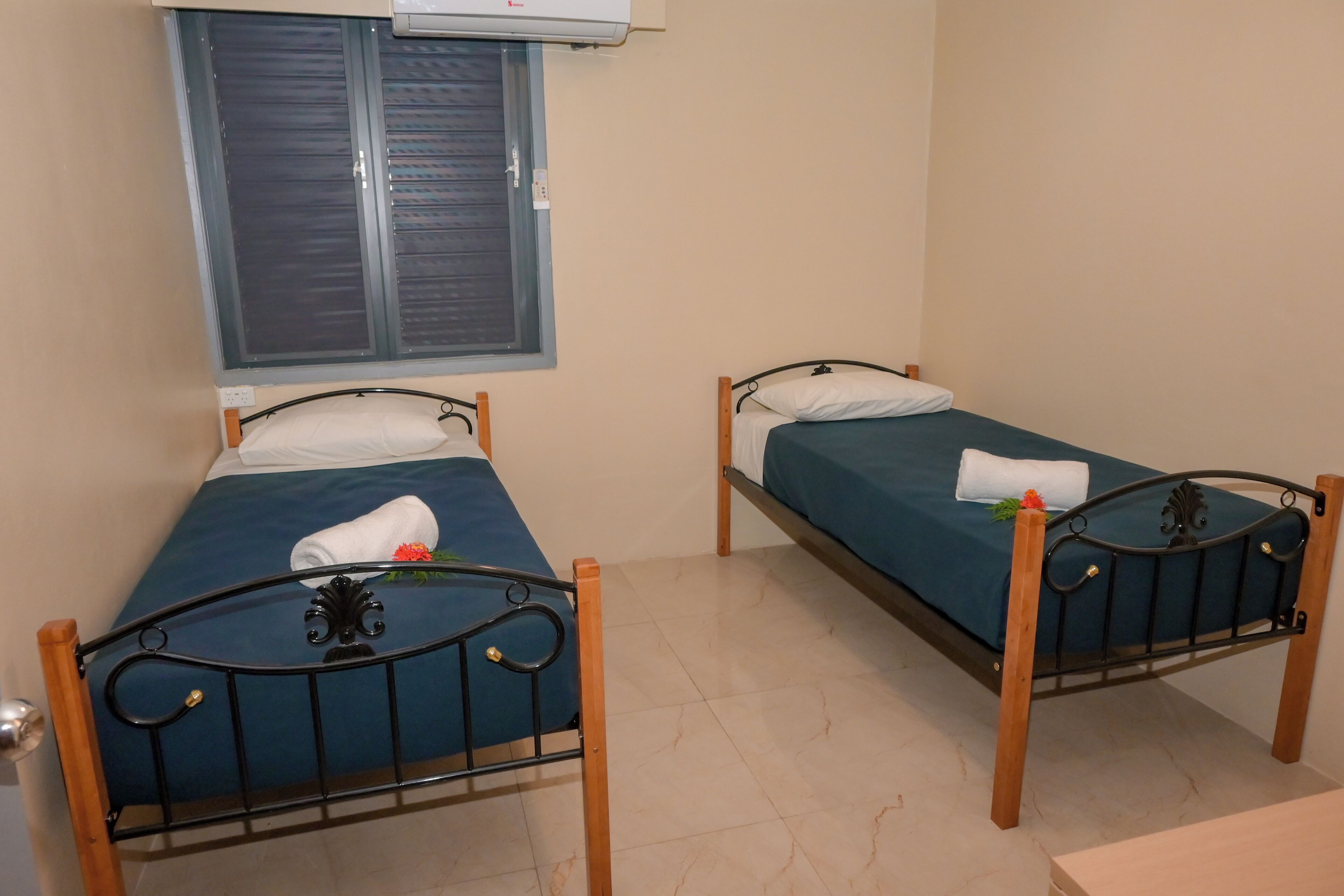 Two single beds with wooden frames and metal headboards, a window with closed shutters, and an air conditioning unit above.