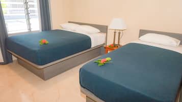 Dos camas juntas con ropa de cama azul a juego y almohadas blancas, una mesita de noche con una lámpara, y una ventana con cortinas.