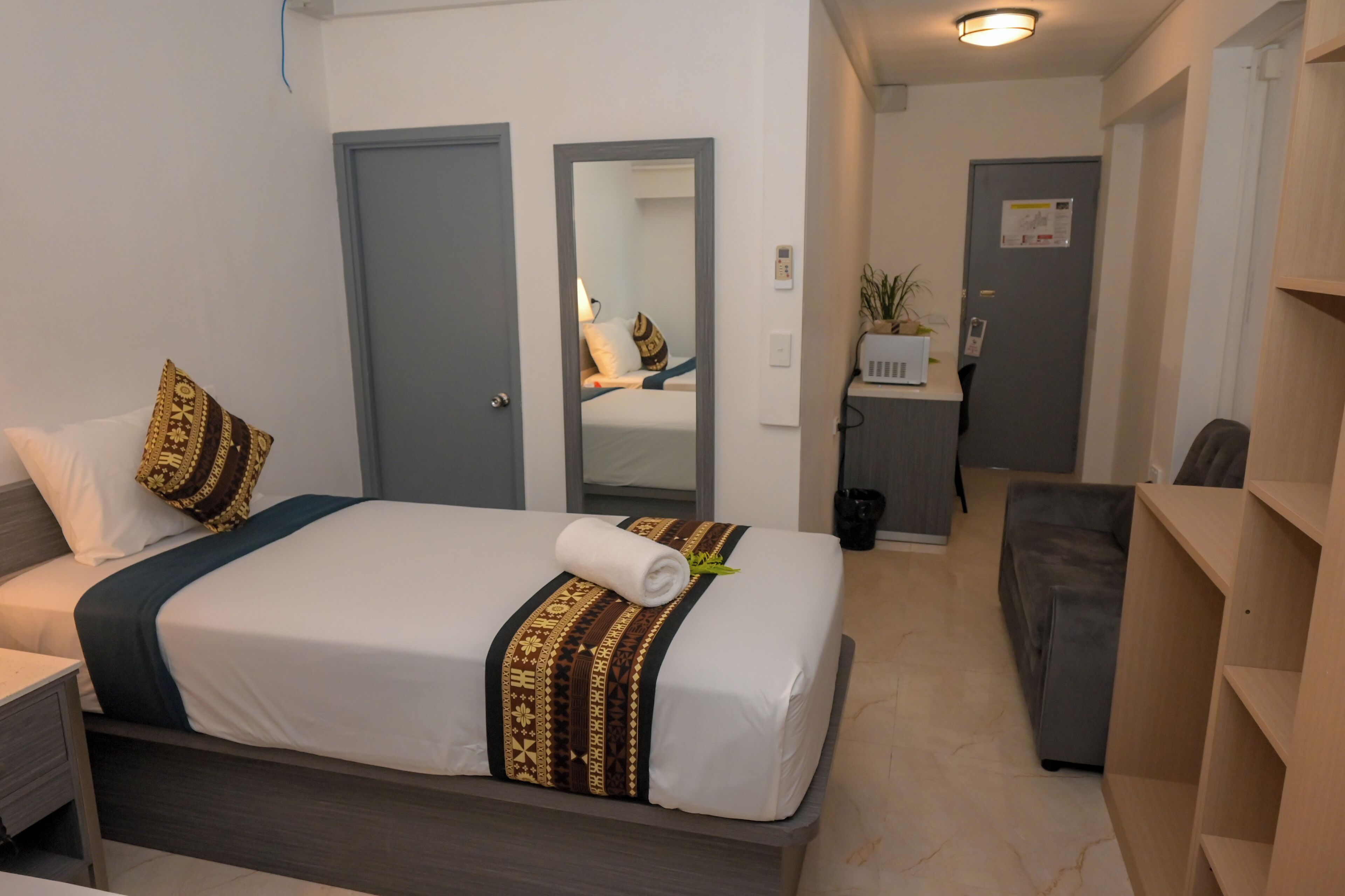 A hotel room with a neatly made bed, a bedside table, a grey sofa, and a small desk with a chair.