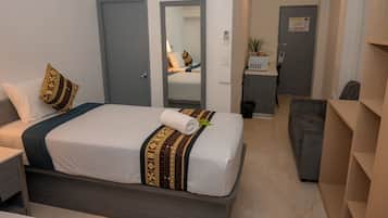 A hotel room with a neatly made bed, a bedside table, a grey sofa, and a small desk with a chair.