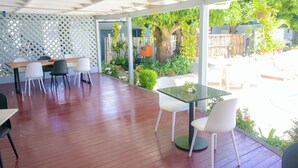 Un patio al aire libre con suelo de madera, sillas y mesas blancas. Cuenta con una pared enrejada y vistas a un jardín con árboles y plantas.