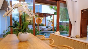 Una mesa de madera para comer con un cuenco blanco de flores, sillas de mimbre y vista a una piscina y zona de estar al aire libre.