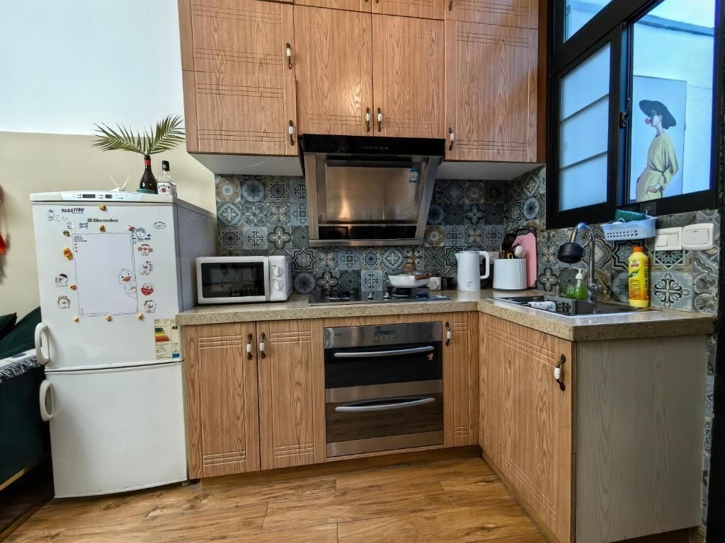 A kitchen with wooden cabinets, a microwave, a refrigerator, and a window with artwork.