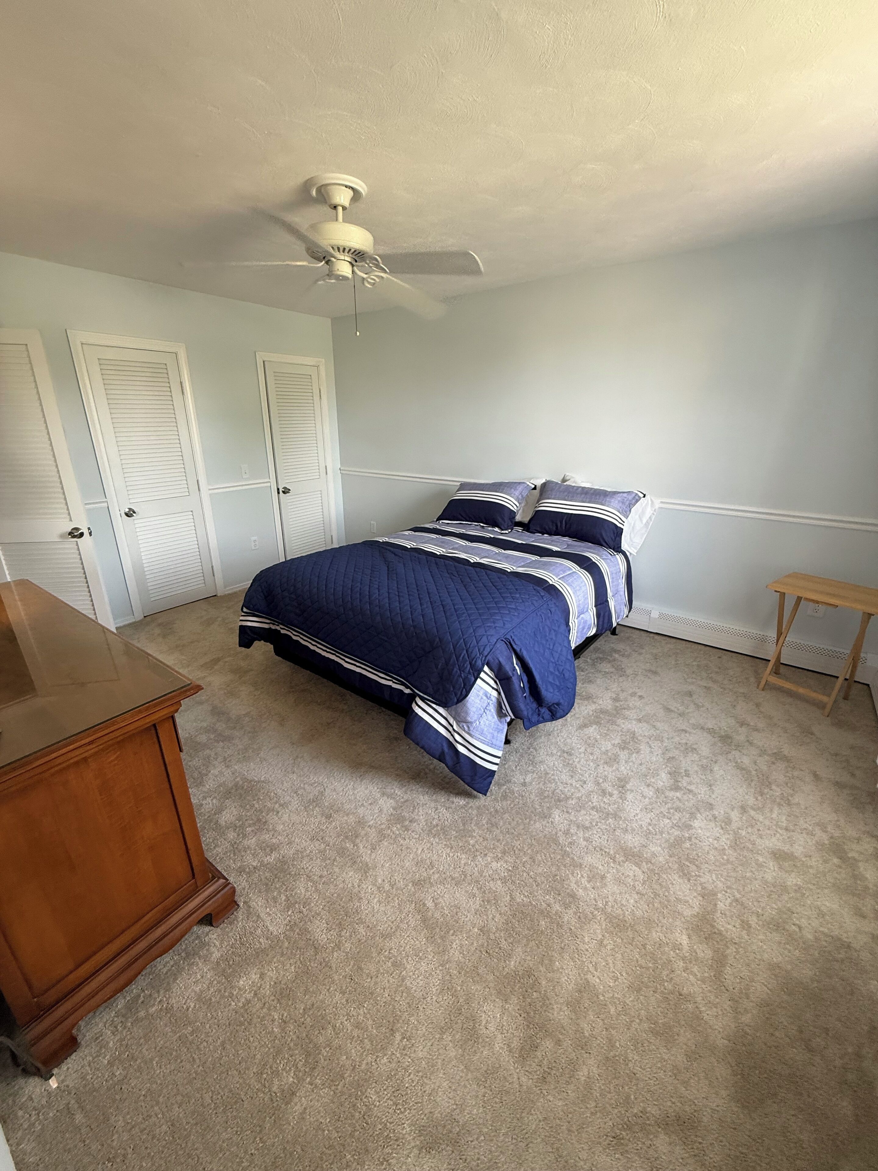 A bedroom with a bed, a ceiling fan, a wooden dresser, and a small wooden table.