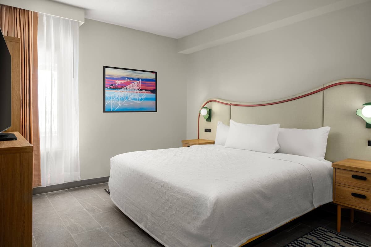 A hotel room with a bed, bedside tables, a TV, and a painting on the wall.