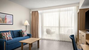 Una camera d'albergo moderna con divano, un tavolo in legno, televisione e un'ampia finestra con tende leggere.