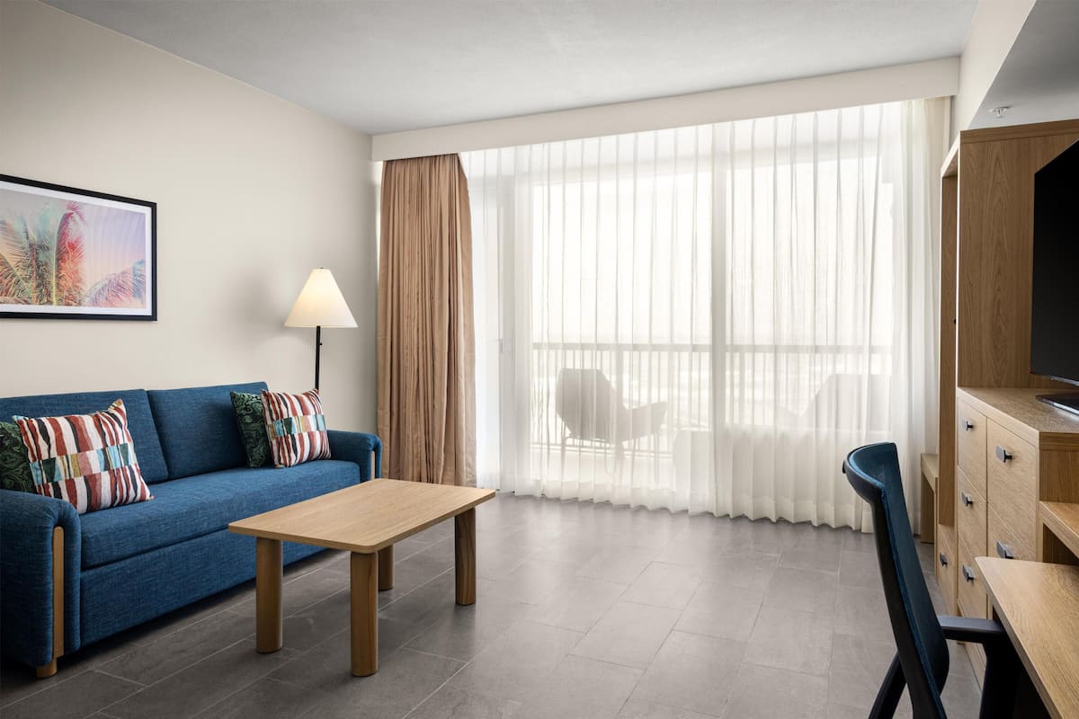 A modern hotel room with a sofa, a wooden table, a TV, and a large window with sheer curtains.