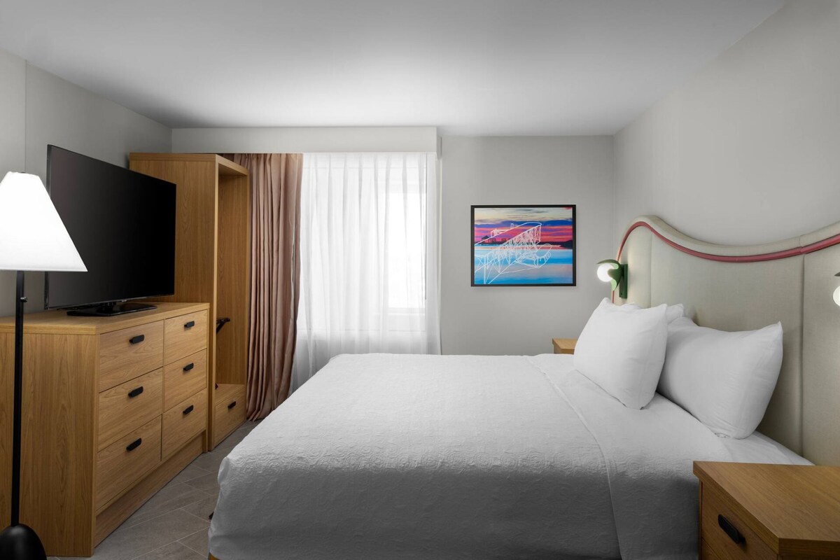 A hotel room with a bed, a TV, a wardrobe, and a painting on the wall.