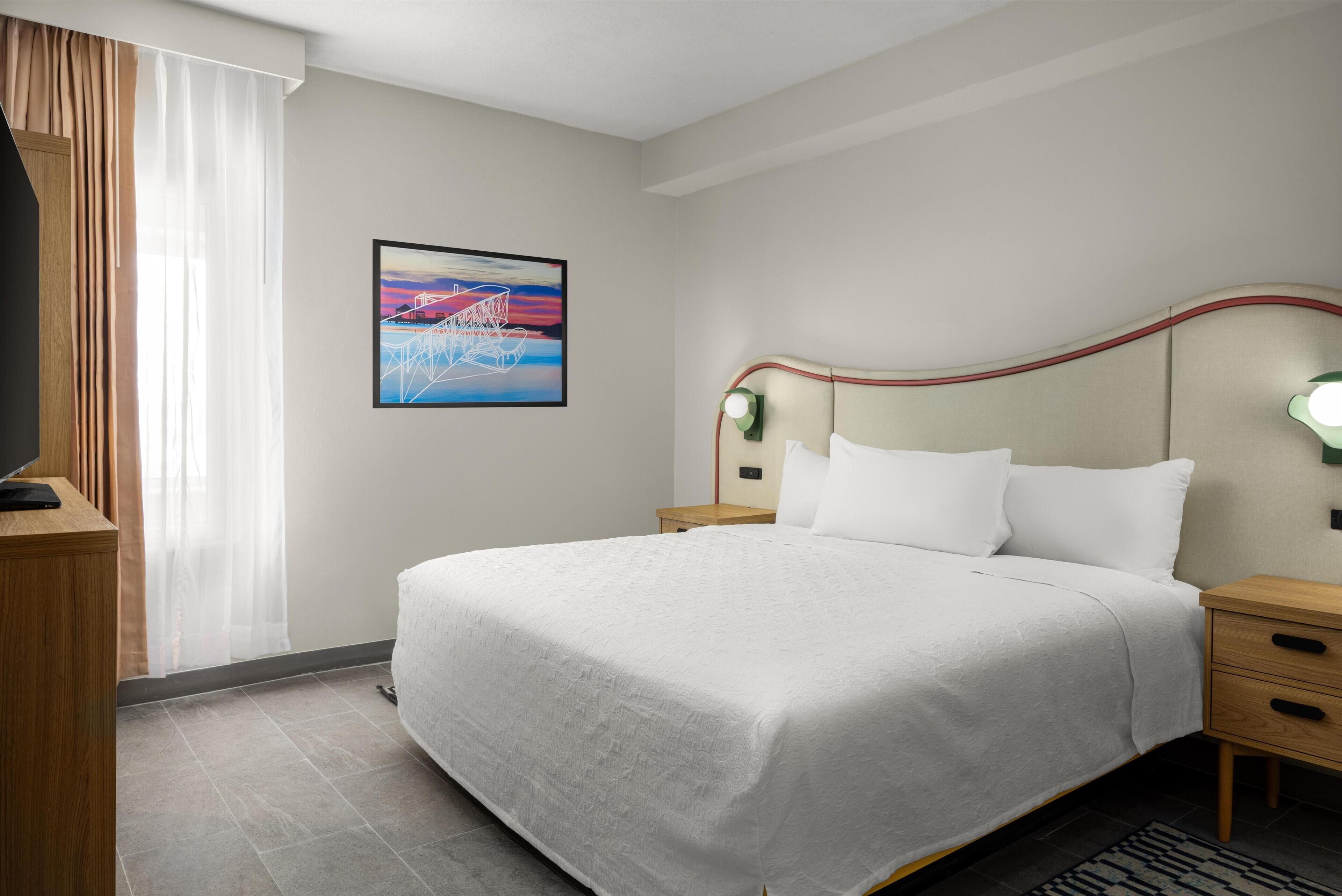 Habitación de hotel con cama, mesita de noche, lámpara, un cuadro y ventana con cortinas.