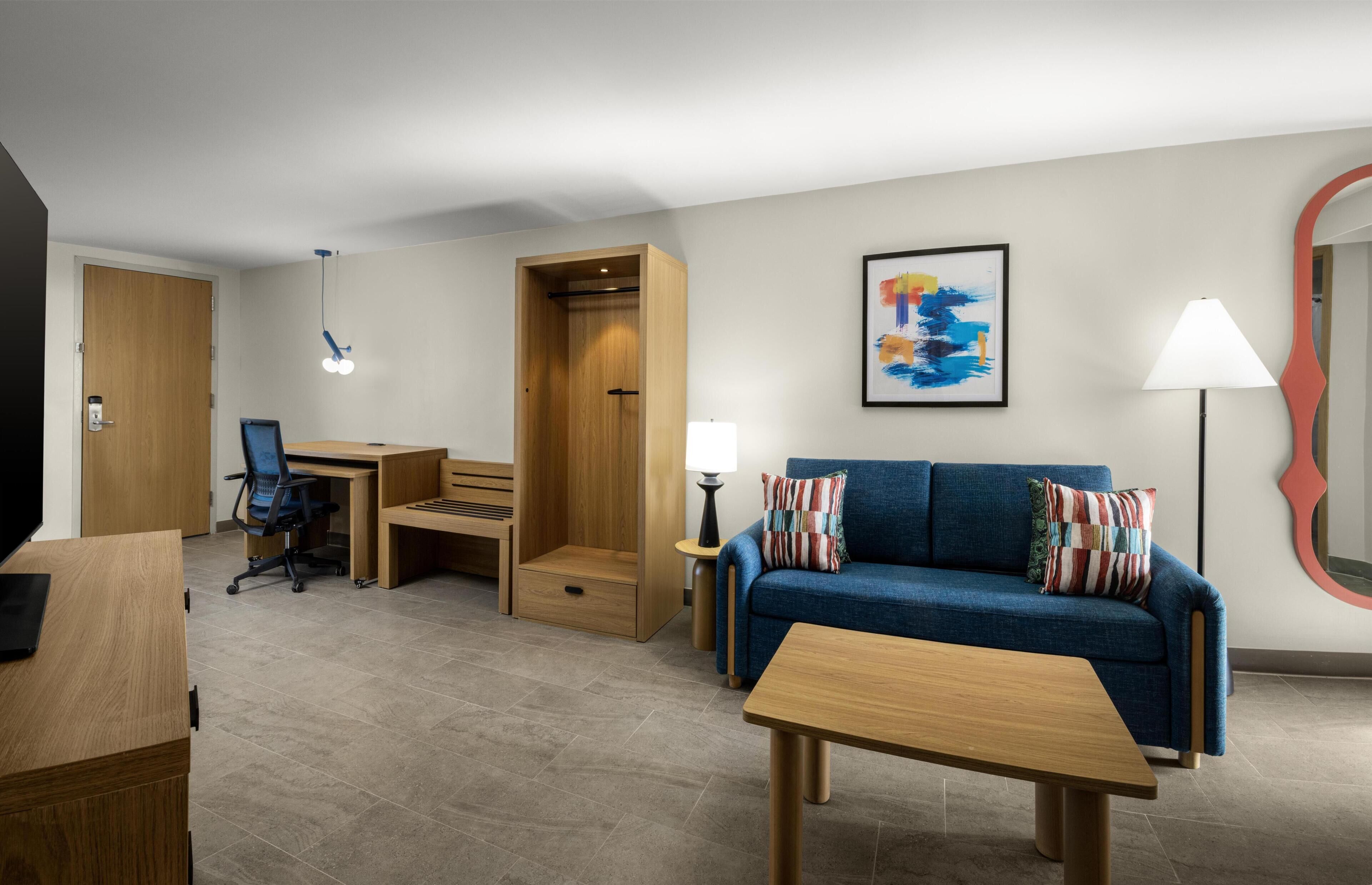 Una camera d'albergo moderna con una scrivania in legno, un divano blu, un tavolino, una lampada e un quadro astratto incorniciato.