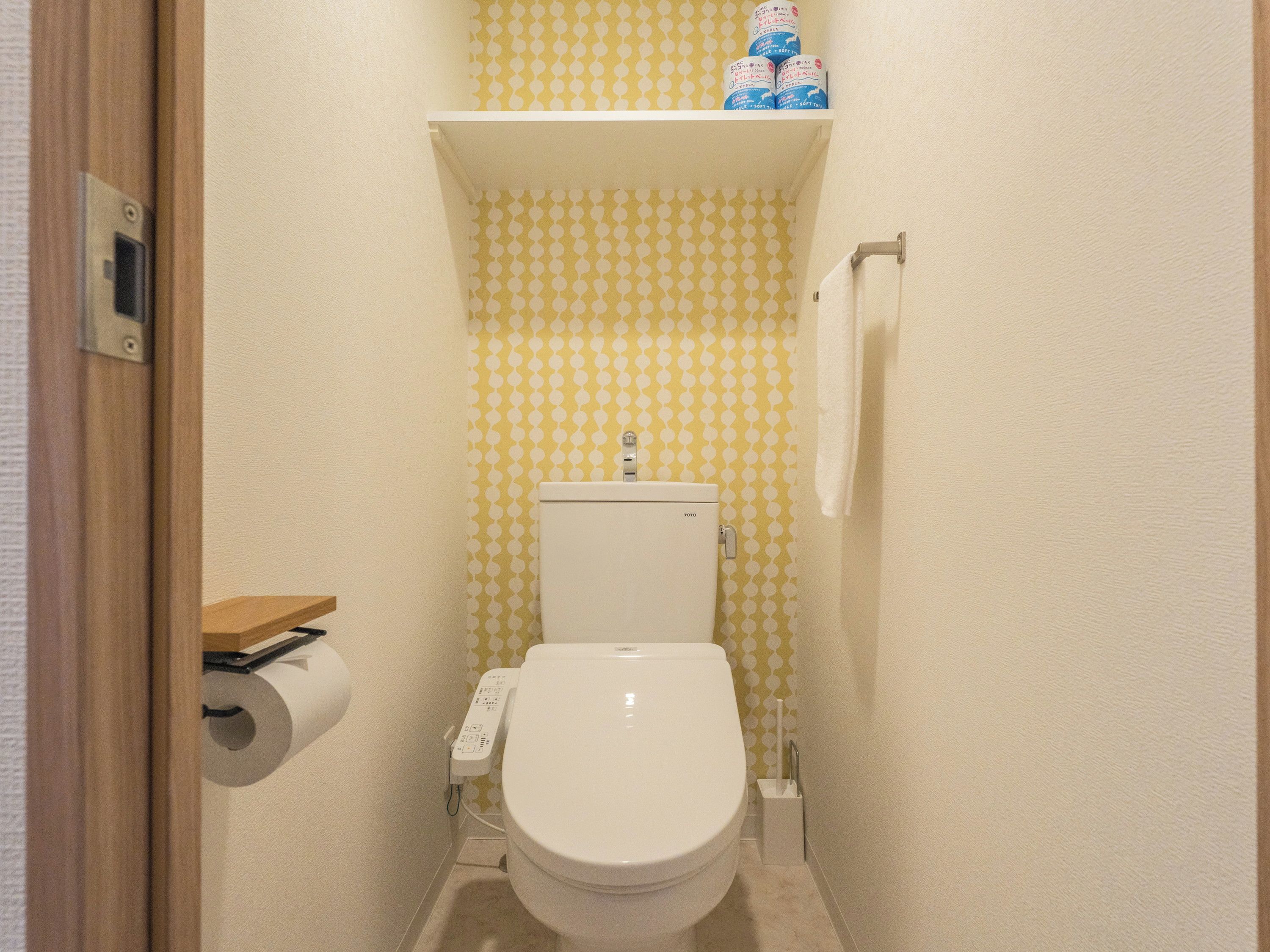 A small bathroom with a toilet, a toilet paper holder, a shelf with toilet paper, and a towel rack.