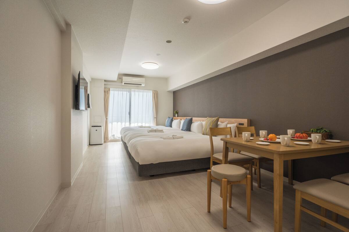 Moderni hotellihuone, jossa on suuri sänky, ruokapöytä tuoleineen, pieni keittiö sekä seinälle asennettu taulutelevisio.