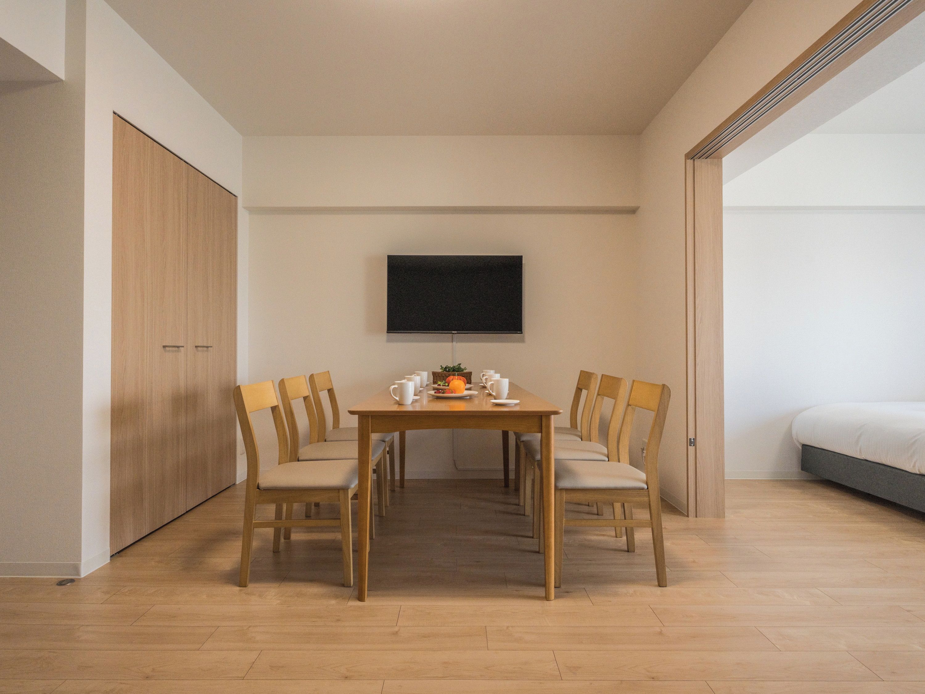 A modern room with a wooden dining table, six chairs, a flat-screen TV mounted on the wall, and a bed with a grey mattress.
