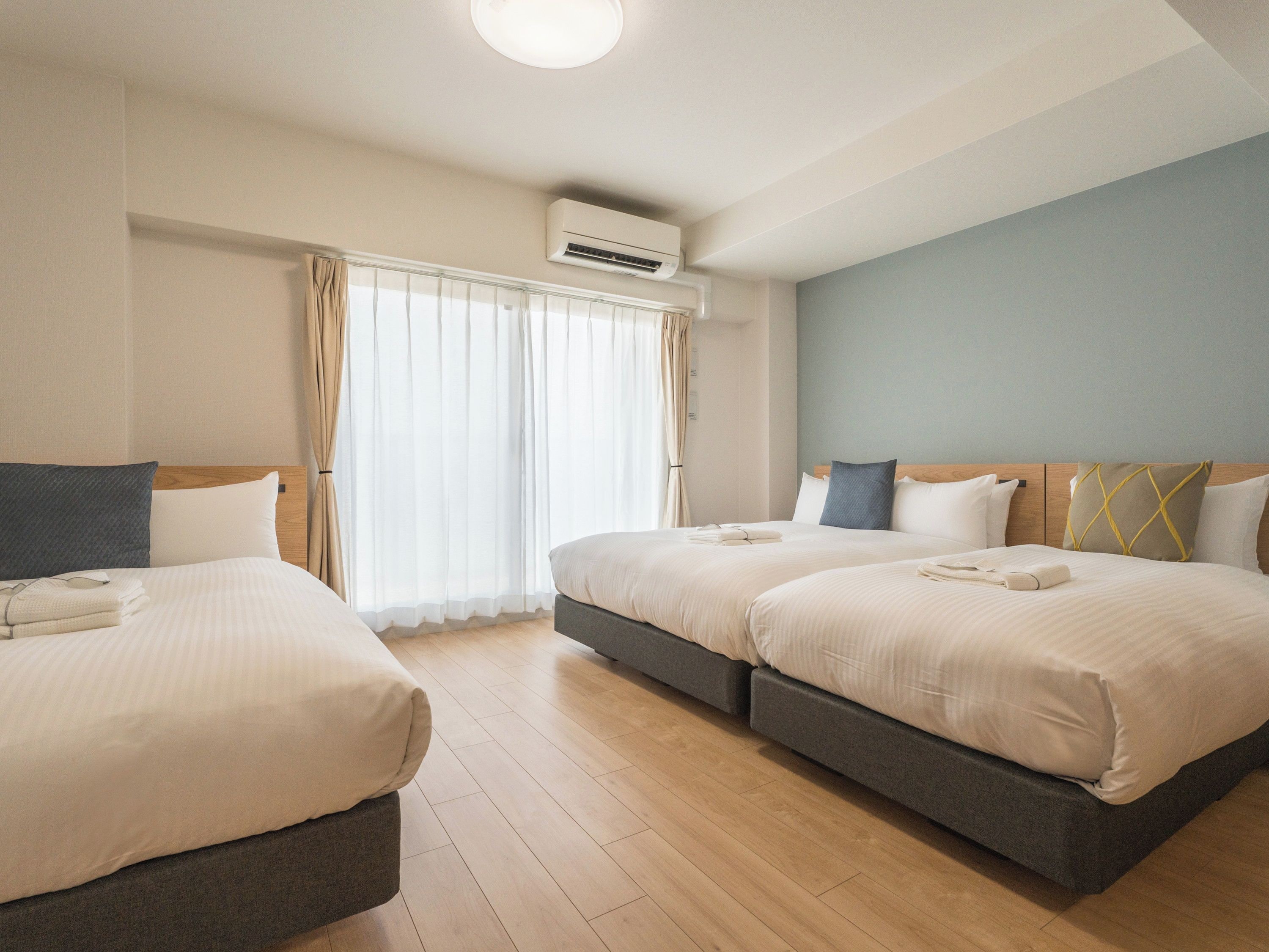 A hotel room with two beds, a window with curtains, an air conditioning unit, and a wooden floor.