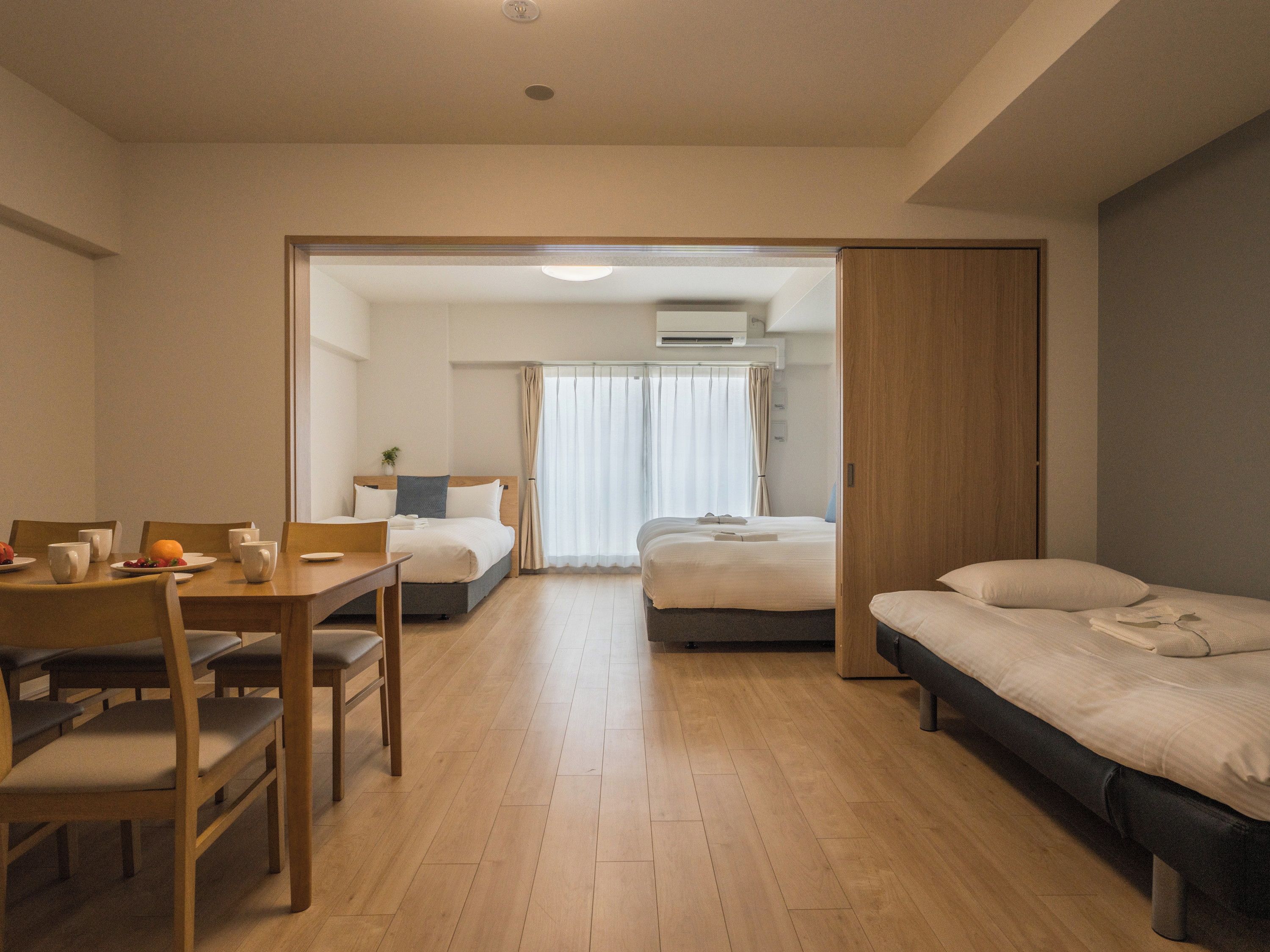 A hotel room with a dining area, two beds, a wooden floor, and a view through an open door.