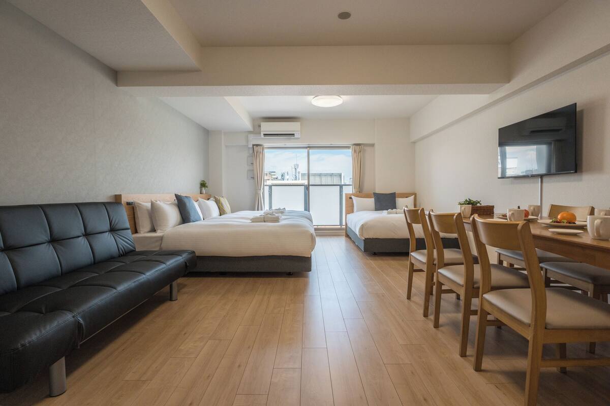 Moderni hotellihuone, jossa on sohva, kaksi sänkyä, ruokapöytä tuoleineen ja seinälle asennettu televisio.