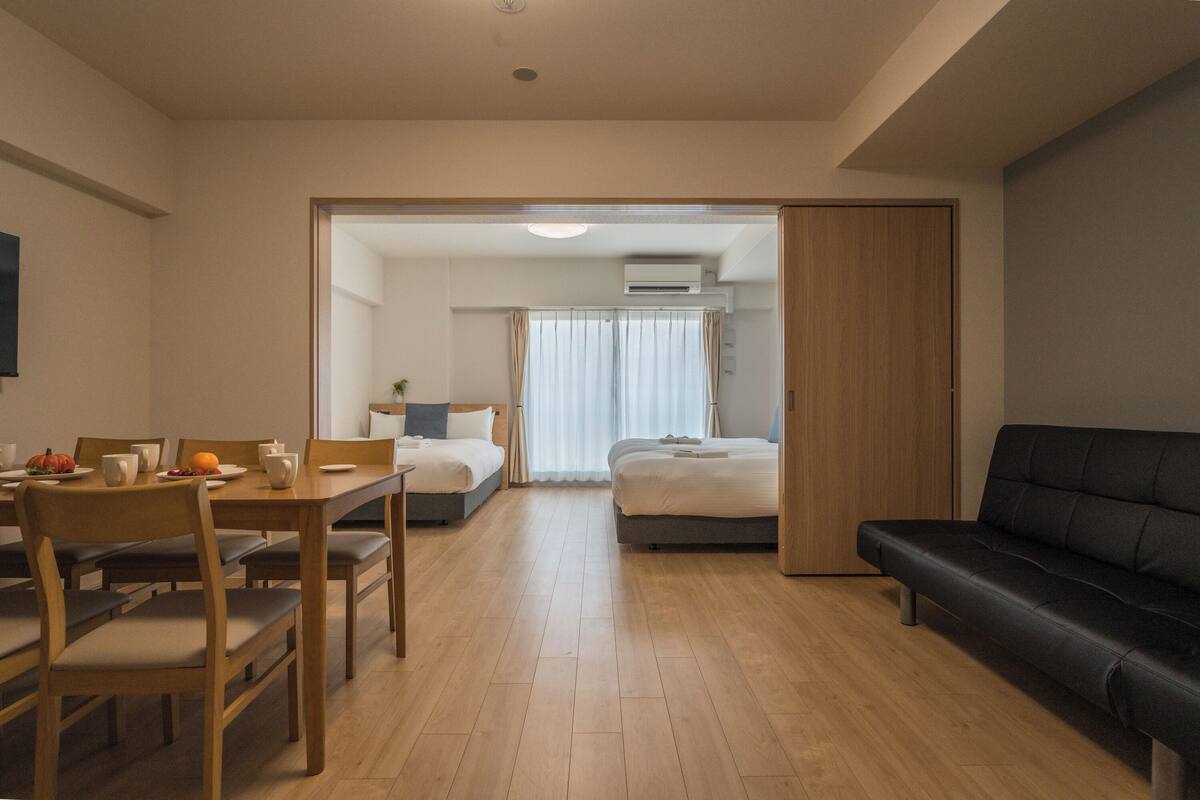 Moderni hotellihuone, jossa on puinen ruokapöytä, musta sohva, sänky ja pieni pöytä, jonka päällä on kasvi.