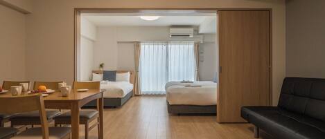 Ein Hotelzimmer mit Holzboden, einem Bett mit weißer Daunendecke, einem schwarzen Sofa, einem Esstisch mit Stühlen und einem kleinen Tisch mit Obstschale und Tassen.
