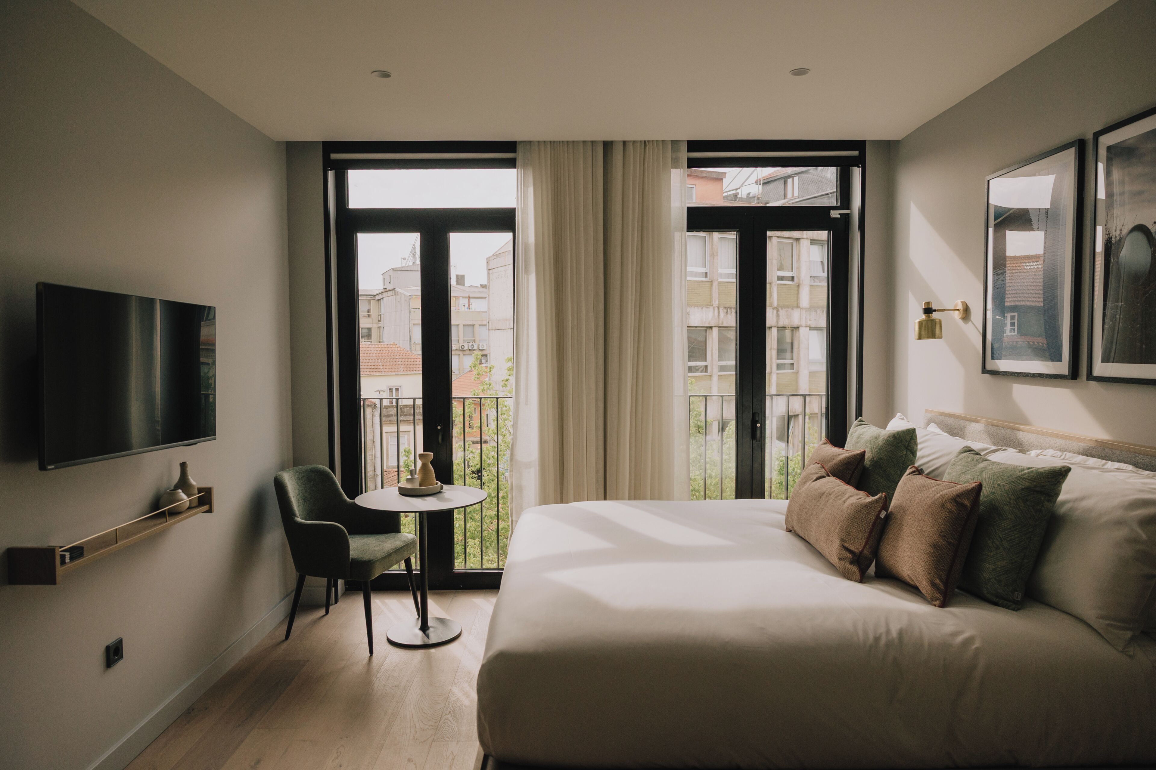 A hotel room with a bed, a TV, a chair, a small table, and a balcony with a view of buildings.