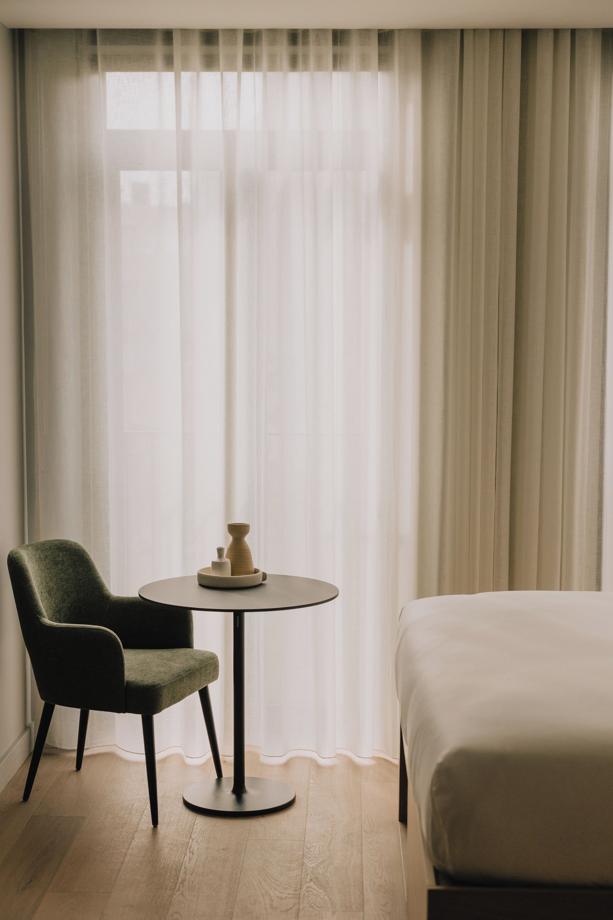 Habitación de hotel con cama, una silla, una pequeña mesa redonda con un florero y una vela, y cortinas de encaje.