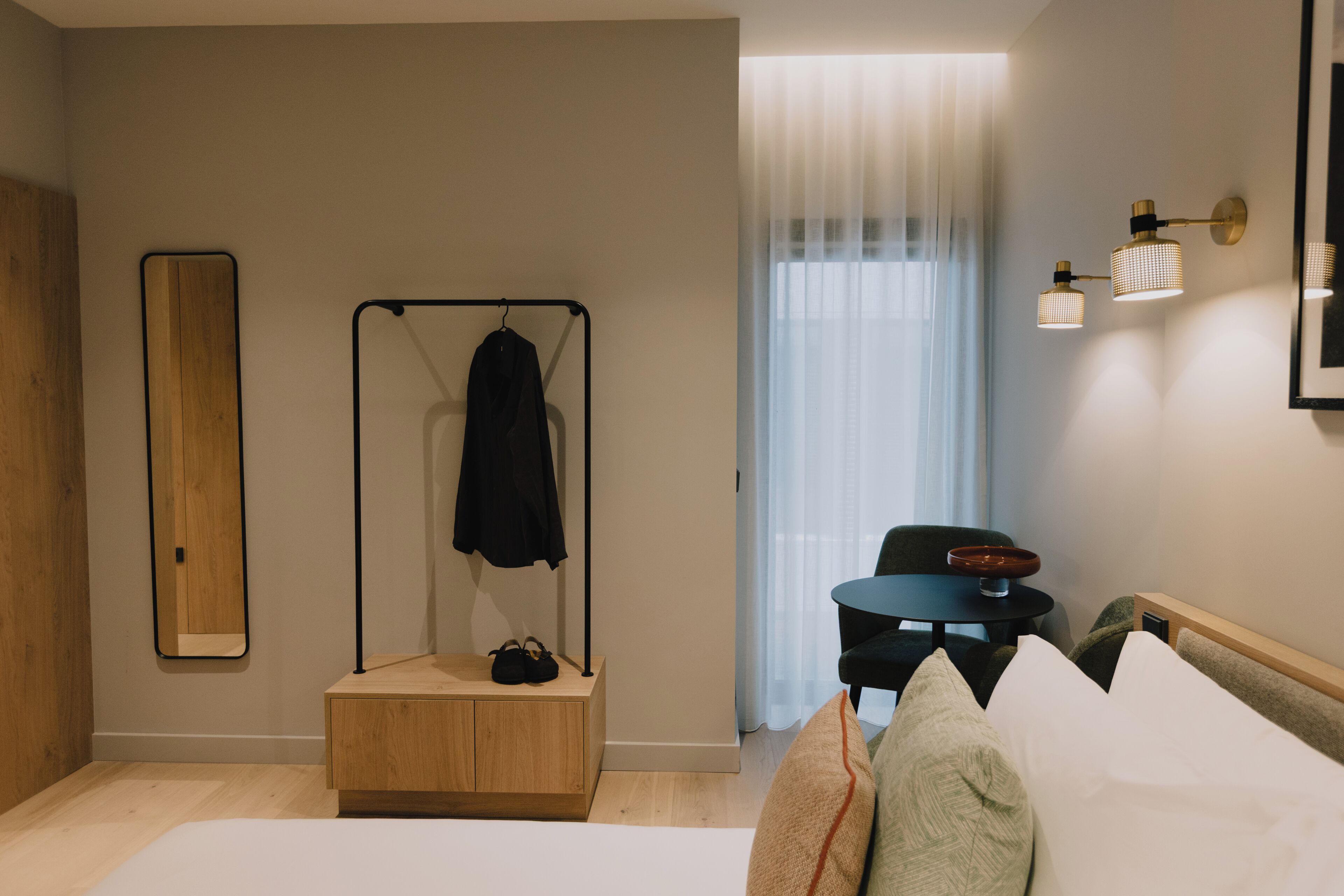 Ein modernes Hotelzimmer mit einem hölzernen Nachttisch, einem schwarzen Kleiderhaken, einem kleinen runden Tisch mit einer Schüssel, einem Stuhl und einem großen Spiegel an der Wand.