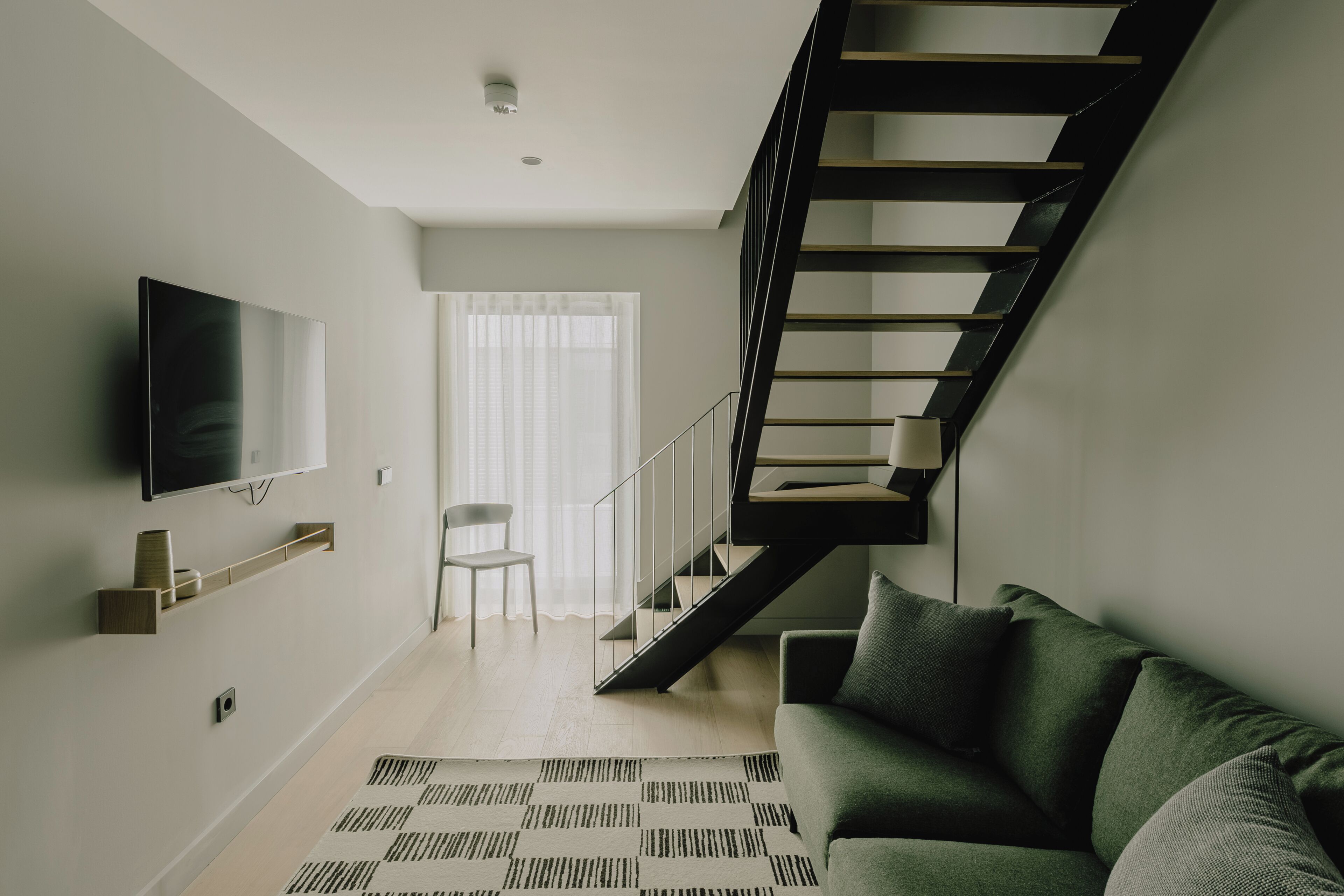 A modern living room with a staircase, a flat-screen TV, a sofa, and a patterned rug.