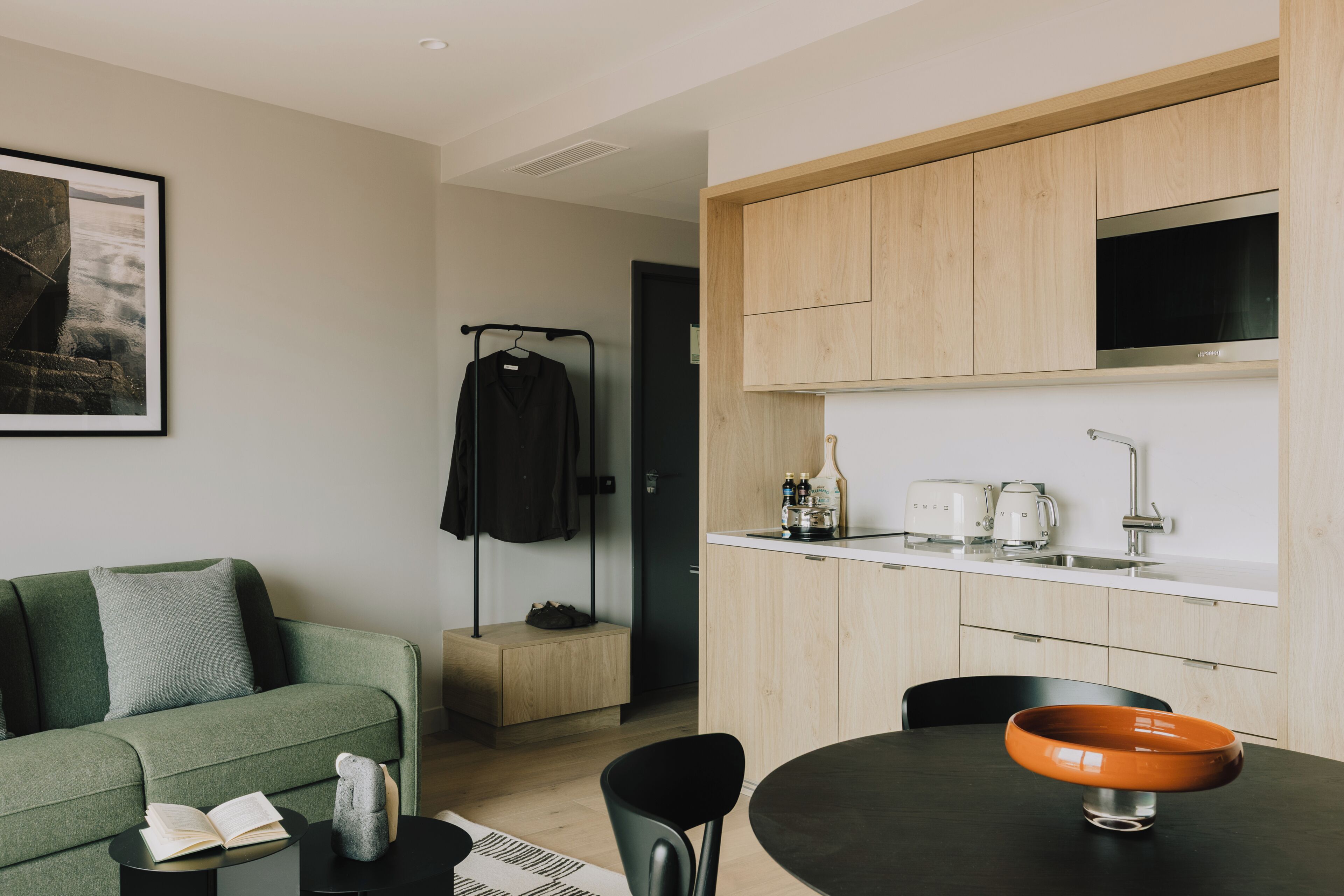 A modern living room with a sofa, a dining table with a bowl, a black chair, a small side table with a book, and a coat rack with a jacket.