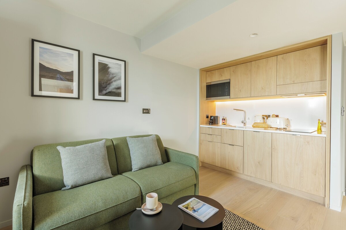 A modern living room with a green sofa, a small round table with a cup and a magazine, and a kitchen area with wooden cabinets and a microwave.