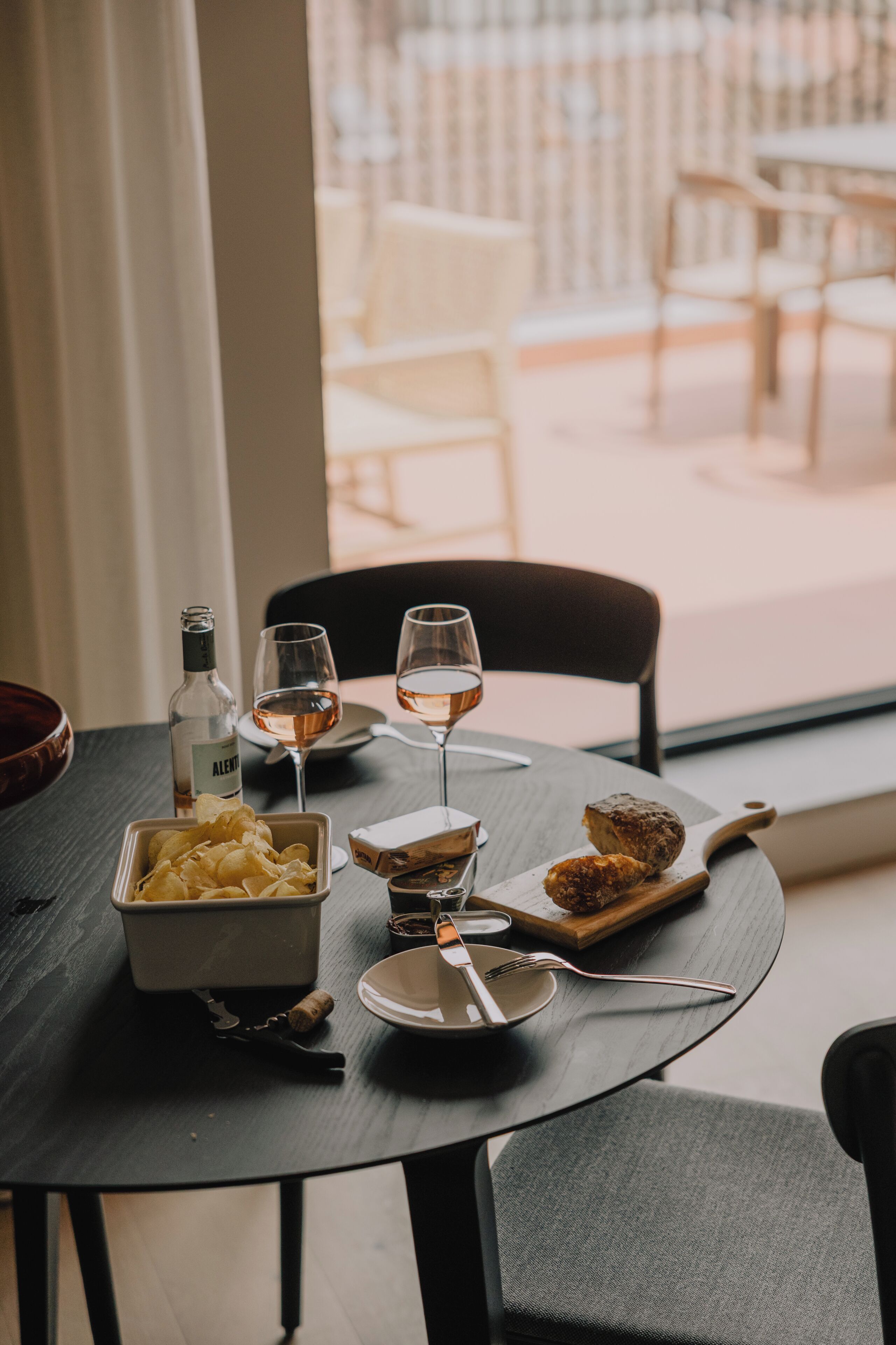 A dining table with a bottle, wine glasses, a bowl of chips, a plate with a pastry, and a wooden board with a pastry.