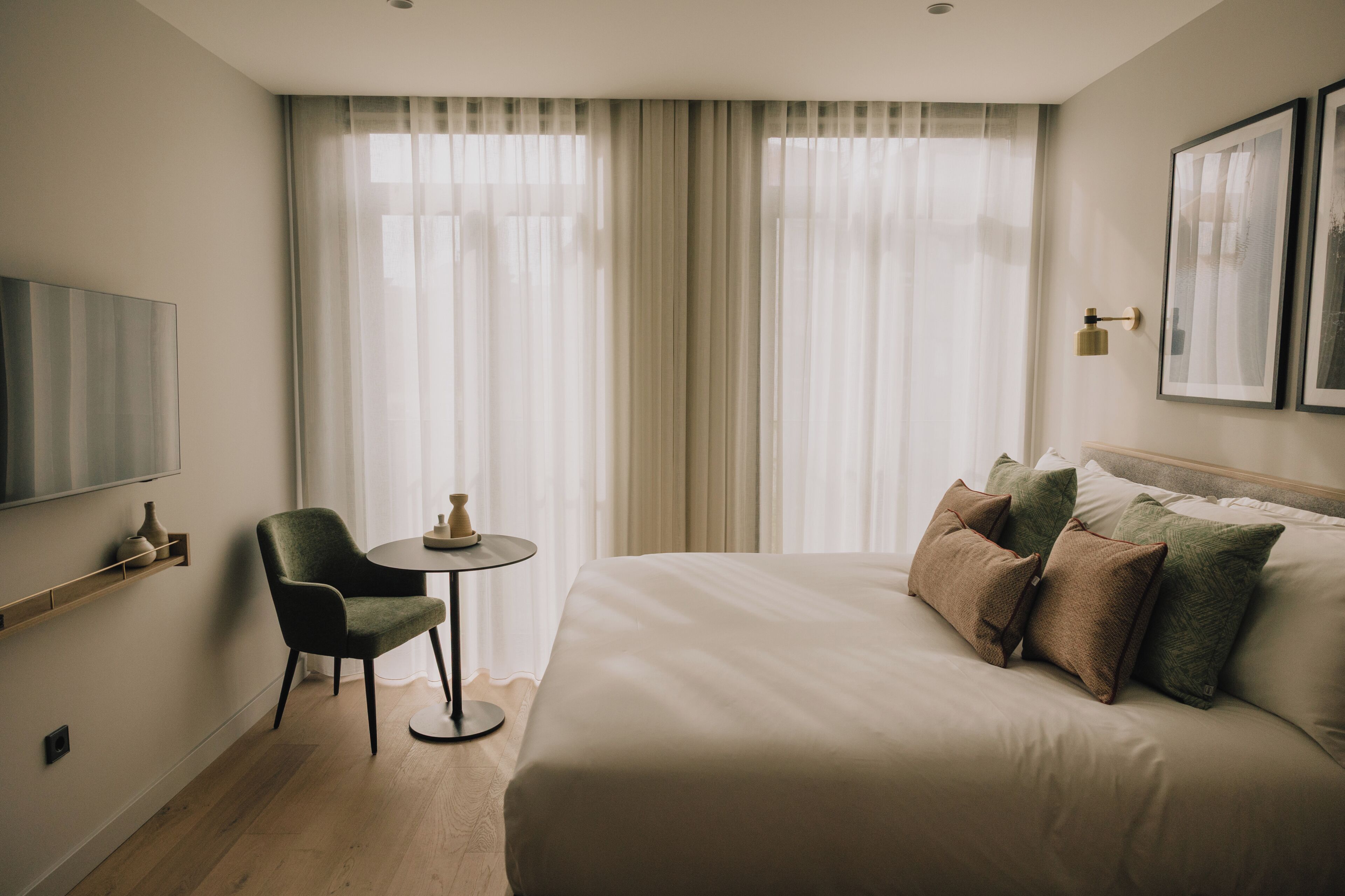 A hotel room with a bed, a chair, a small table, and a TV on the wall.