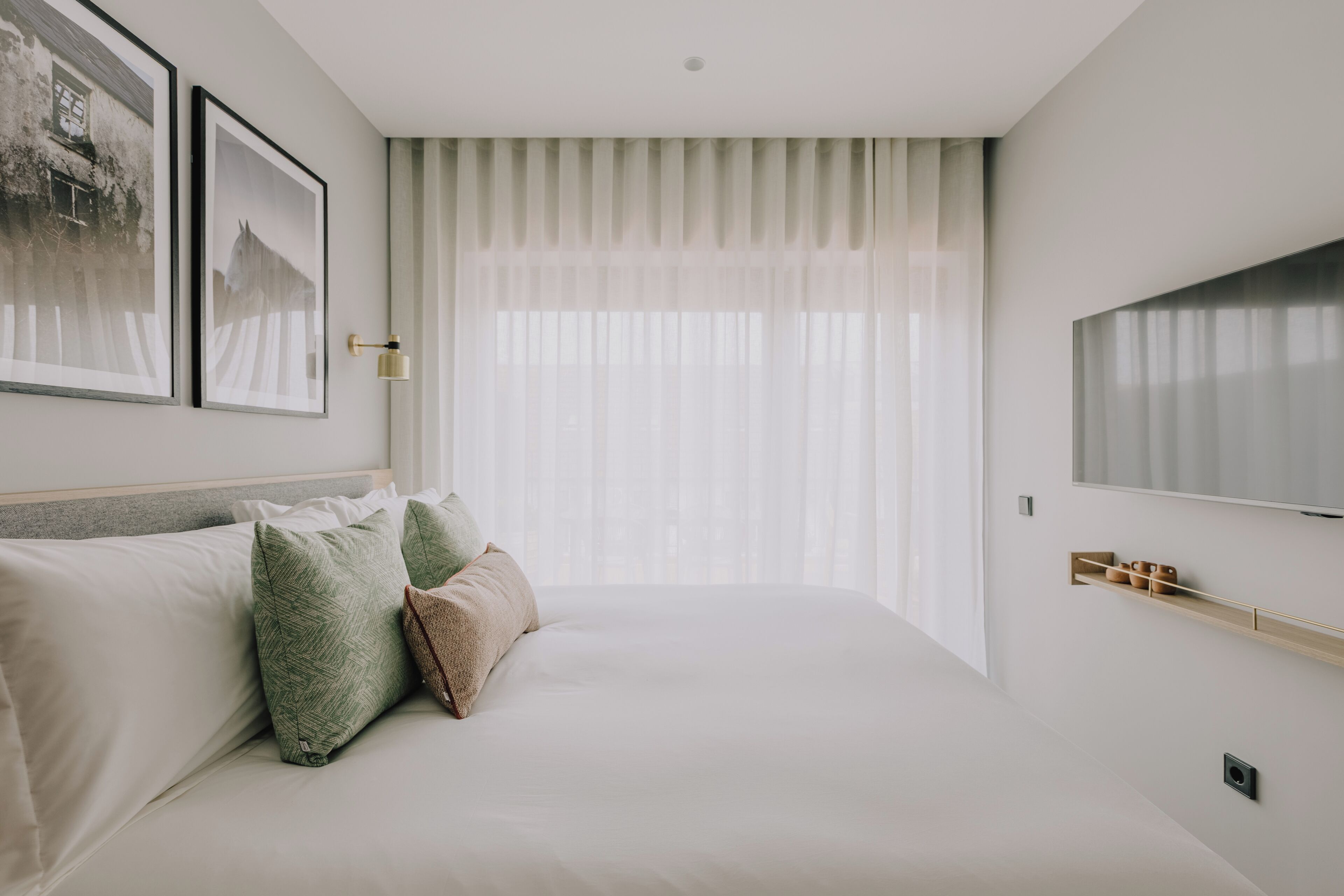 A modern bedroom with a large bed, a flat-screen TV, and a window with sheer curtains.