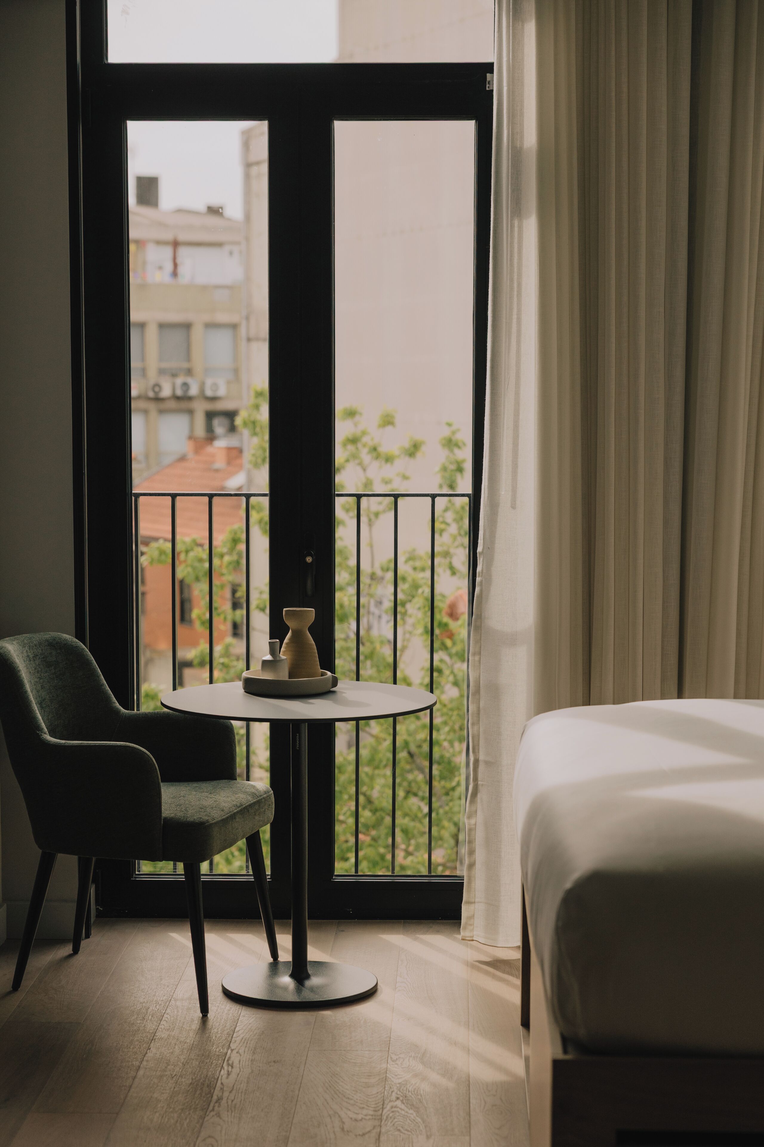 A hotel room with a bed, a chair, a small table, and a view of buildings through a window.