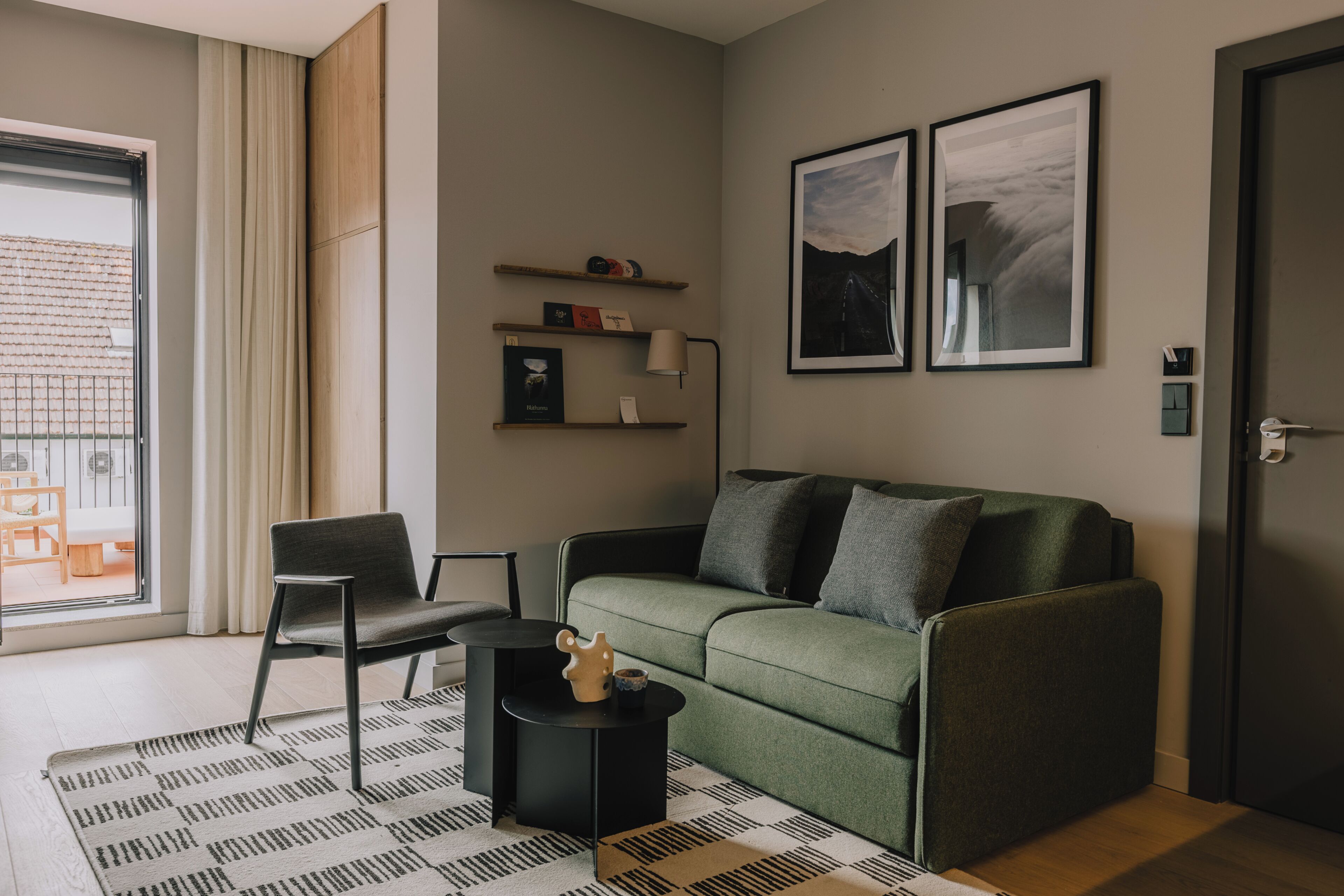 En moderne stue med en grøn sofa, en sort lænestol, et lille sofabord og en gulvtæppe med et mønster. To billeder hænger på væggen, et vindue med gardiner og en dør til højre.