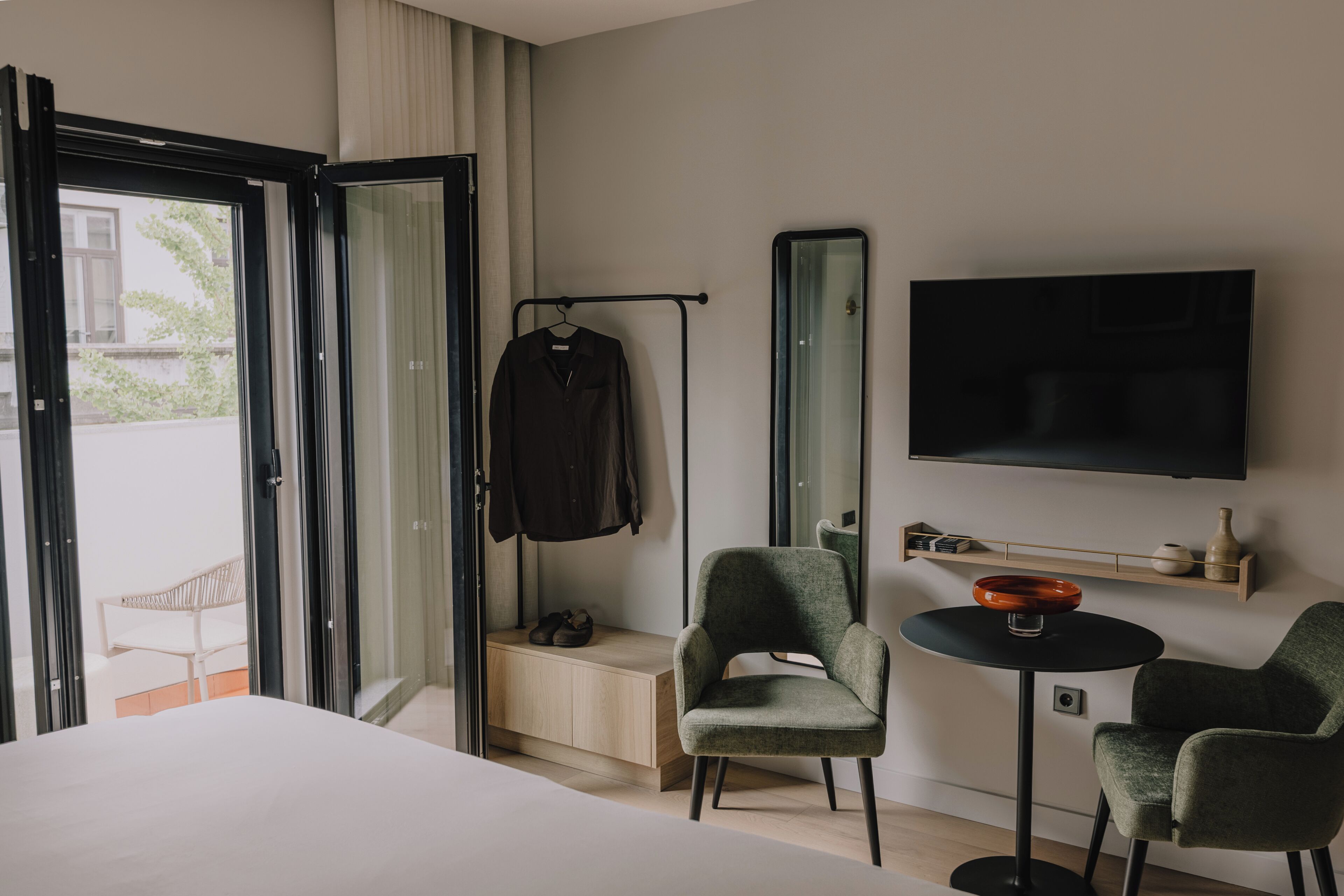 A hotel room with a bed, a seating area with two chairs, a small table, a TV, a coat rack, and a window with a view of the outside.