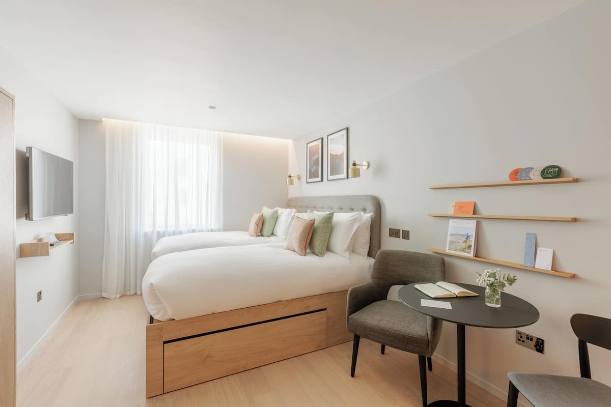 A modern hotel room with a large bed, a desk, and a chair. There are shelves with books and decorative items on the wall.