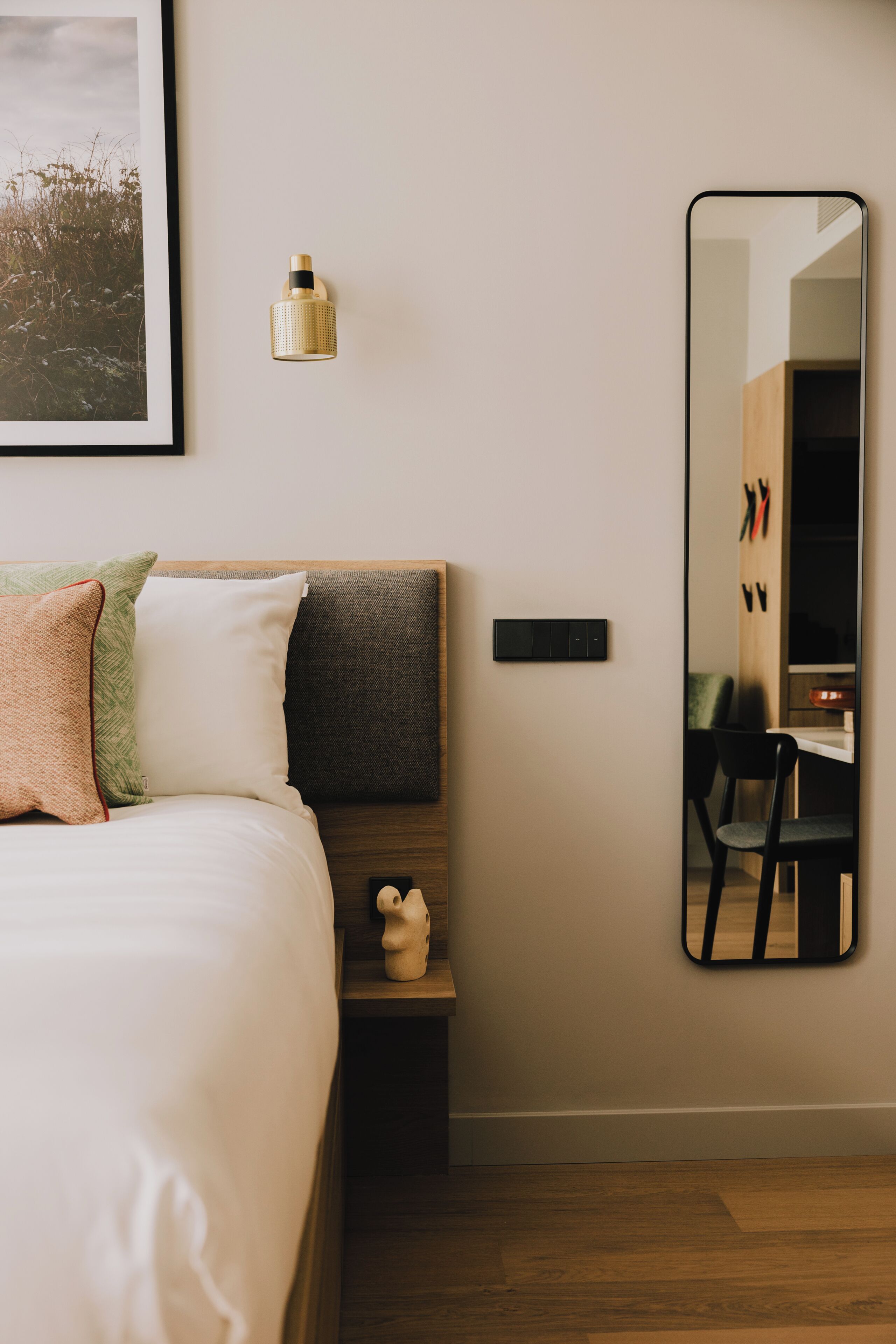 A bedroom with a bed, a nightstand, a mirror, and a wall-mounted light fixture.