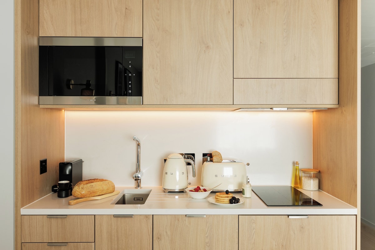A modern kitchen with wooden cabinets, a built-in microwave, a coffee maker, a toaster, a bread loaf, a bowl of fruit, and a bottle of olive oil.