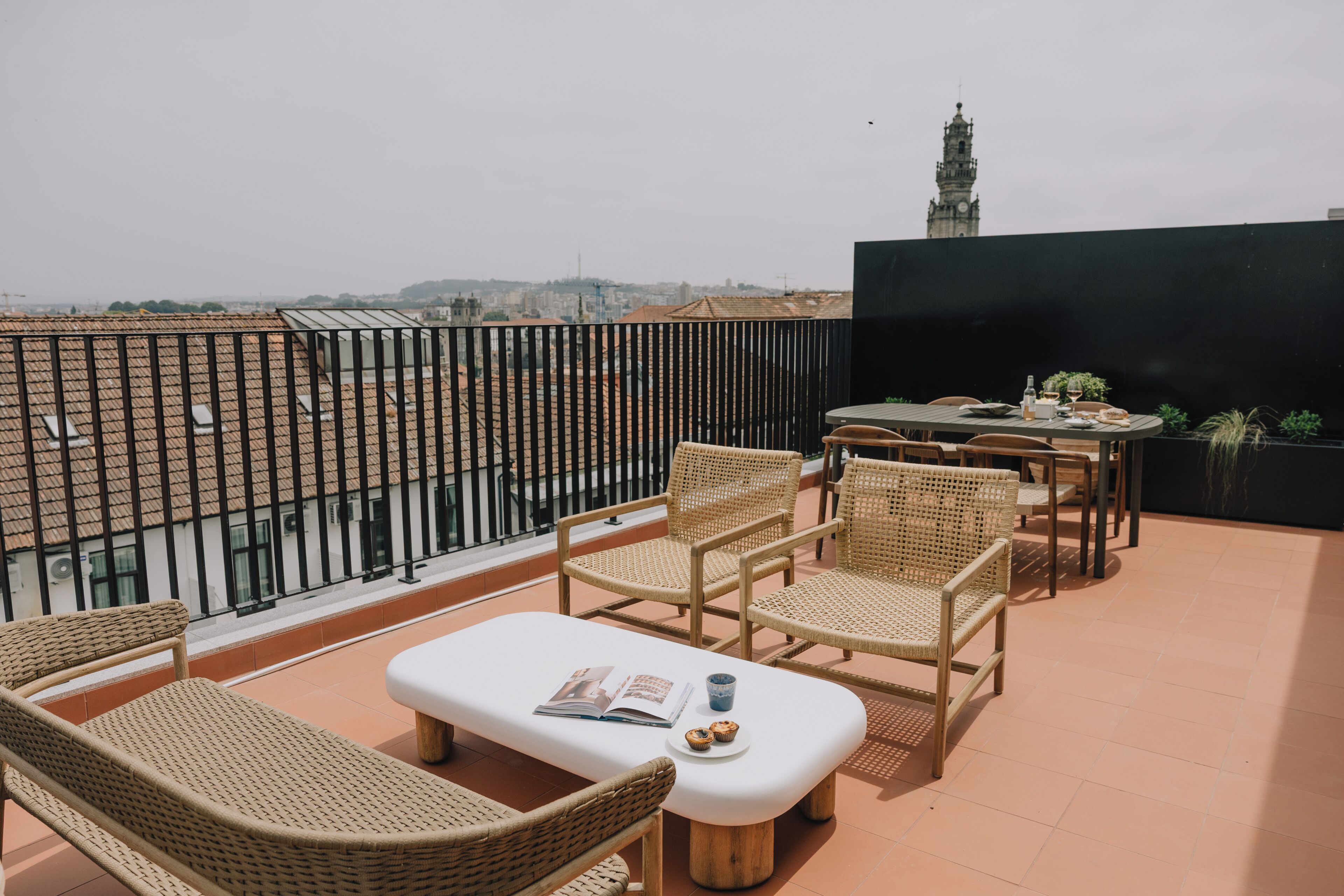 A rooftop terrace with wicker furniture, a table with a drink, and a view of a cityscape and a tower.