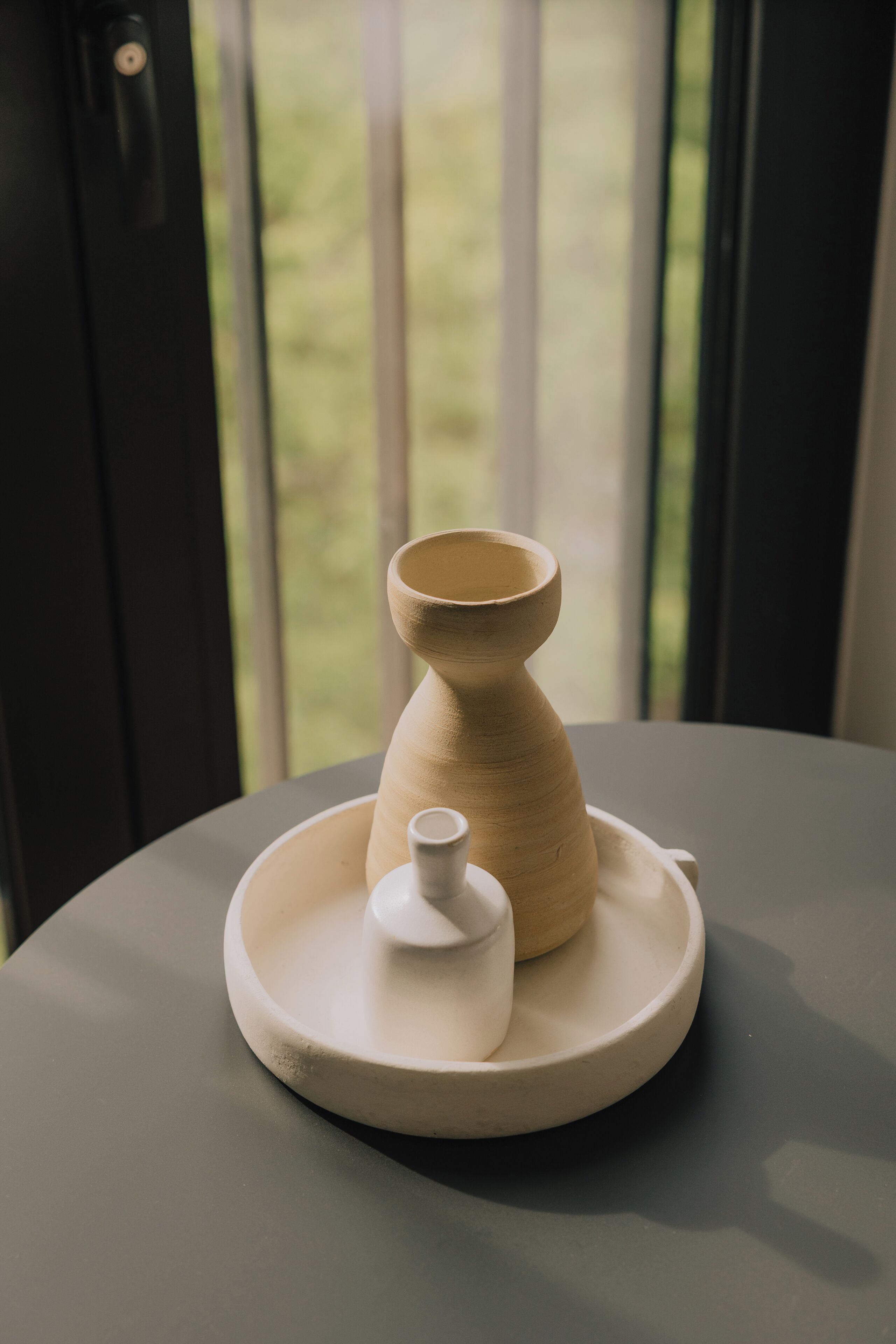 A table with a ceramic vase and a small container on a plate, set against a window with a view of greenery.