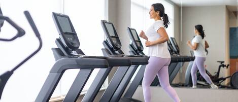 A woman running on a treadmill in a gym with multiple treadmills and a large mirror reflecting the scene.